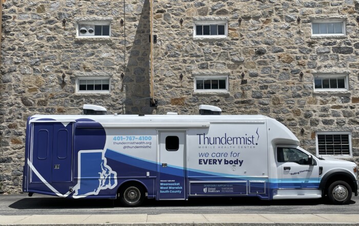 A view of Thuhndermist's mobile medical unit parked outside the West Warwick health center, which is a converted cotton shed featuring a tall stone wall.