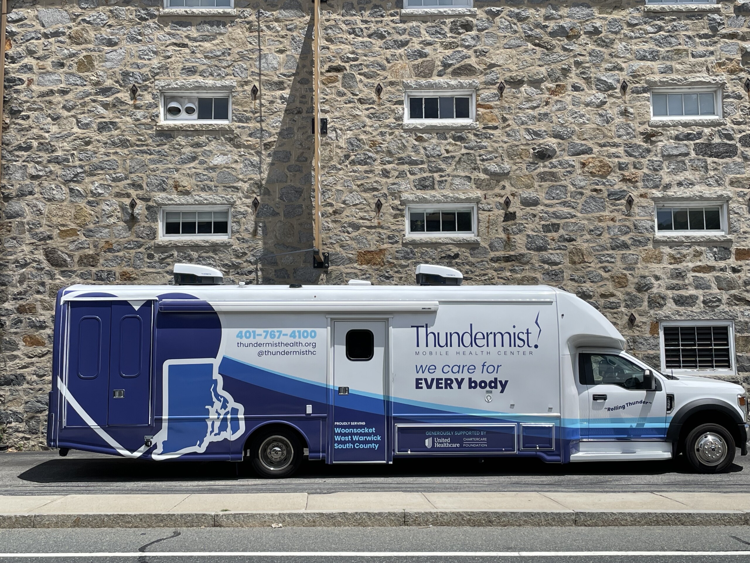 A view of Thuhndermist's mobile medical unit parked outside the West Warwick health center, which is a converted cotton shed featuring a tall stone wall.